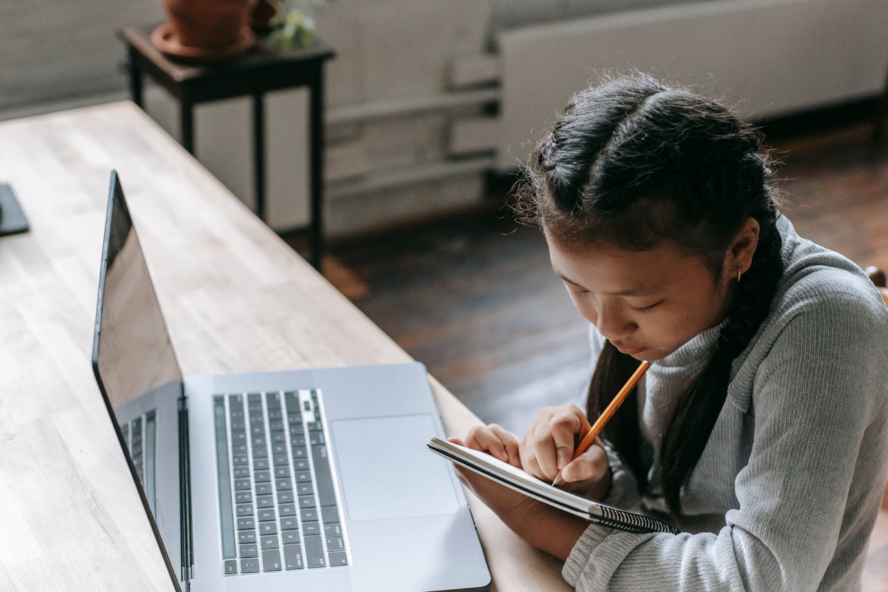 gallery-1 High angle of Asian girl with pigtails writing notes in notepad while studying at desk with laptop